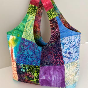 Puede incluir: Bolso tote patchwork colorido con una variedad de cuadrados de tela batik vibrantes. El bolso presenta un asa curva y una mezcla de azules, verdes, morados y rosas. El bolso está diseñado con una construcción acolchada.