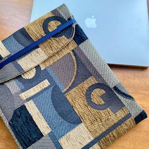 Puede incluir: Una funda para portátil estampada con un diseño geométrico en tonos azules, marrones y grises. La funda tiene un cierre de solapa asegurado por una banda elástica azul. Un portátil plateado está en el fondo.