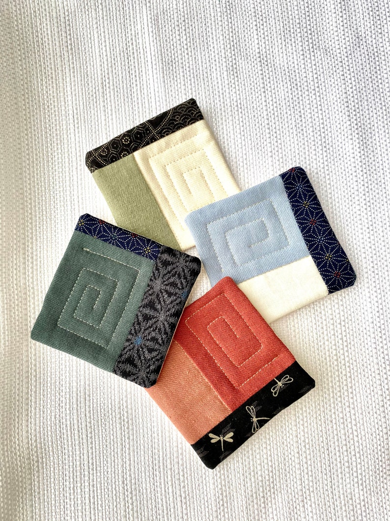Pode incluir: Um conjunto de cinco bases quadradas com um design de espiral acolchoada. Cada base apresenta uma combina&ccedil;&atilde;o de cores &uacute;nica, incluindo verde, azul, creme e ferrugem, com detalhes em tecido estampado. As bases est&atilde;o dispostas sobre uma superf&iacute;cie branca texturizada.