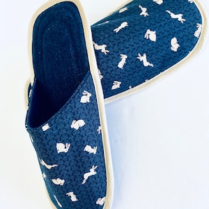 Pu&ograve; includere: Un paio di pantofole blu scuro con un motivo di conigli bianchi. Le pantofole hanno un bordo color crema intorno ai bordi.