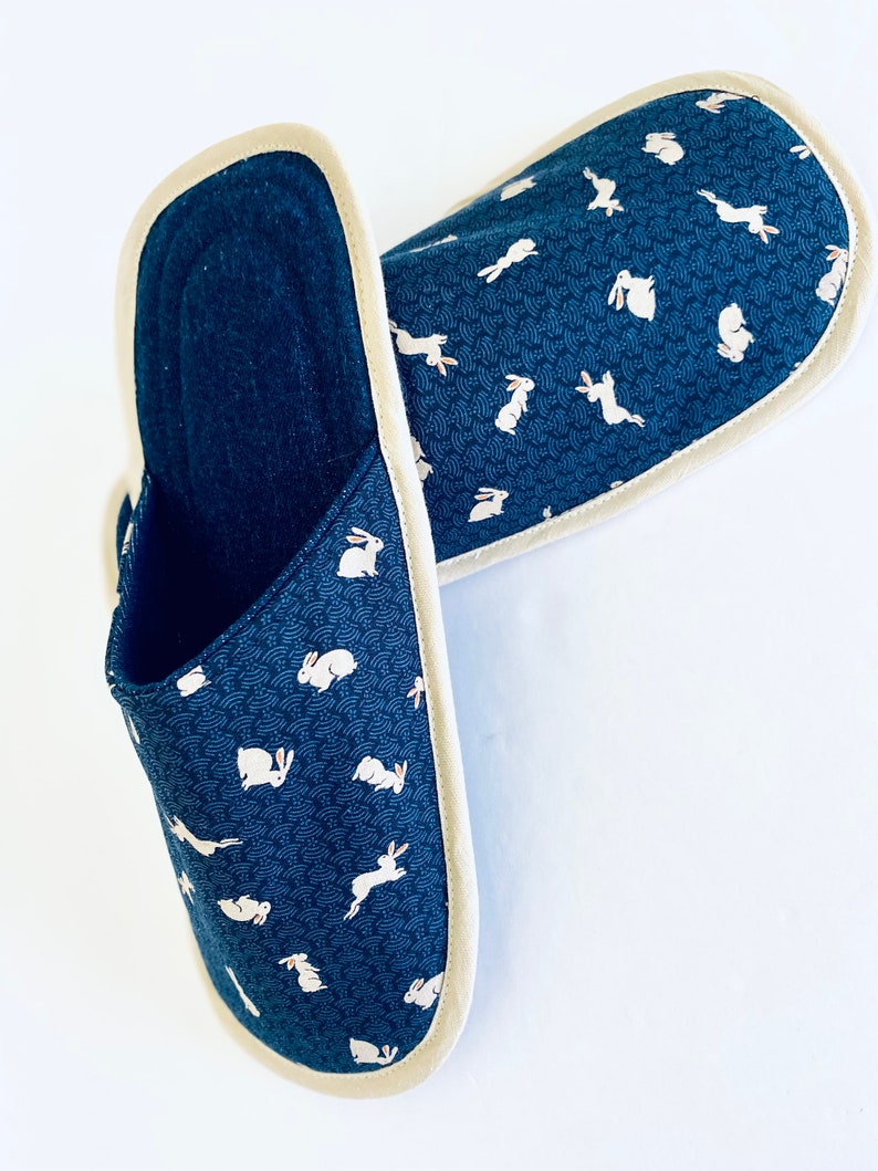 Pu&ograve; includere: Un paio di pantofole blu scuro con un motivo di conigli bianchi. Le pantofole hanno un bordo color crema intorno ai bordi.