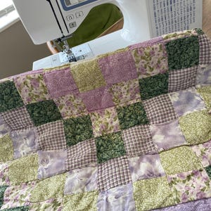 May include: A patchwork quilt in shades of green, lavender, and white is being sewn on a Brother sewing machine. The quilt features a variety of floral and plaid patterns. The machine is white and blue.
