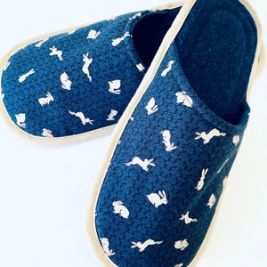 Pu&ograve; includere: Un paio di pantofole blu e bianche con motivo e bordatura color crema. Le pantofole presentano un motivo ripetitivo di conigli bianchi su uno sfondo blu.