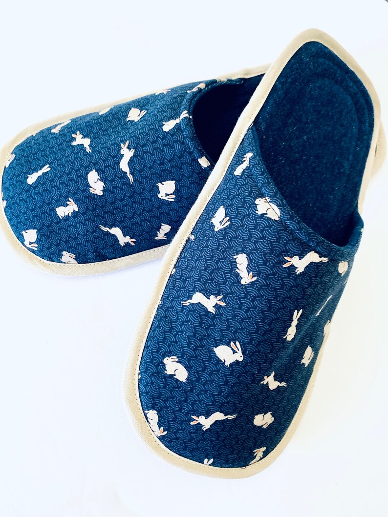 Pu&ograve; includere: Un paio di pantofole blu e bianche con motivo e bordatura color crema. Le pantofole presentano un motivo ripetitivo di conigli bianchi su uno sfondo blu.