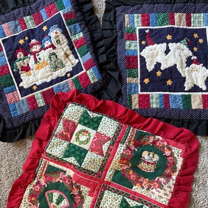 May include: Three square quilted pillows with ruffled edges. Two pillows feature dark blue centers with appliqued snowmen and Santa figures, surrounded by multi-colored patchwork borders. One pillow has a red ruffled edge and four panels.