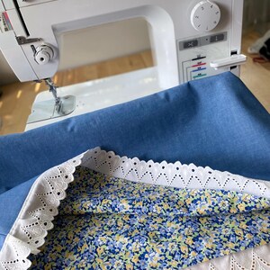 Pode incluir: Uma máquina de costura Brother PQ1500SL branca com tecido azul e um tecido com padrão floral com acabamento em renda vazada branca. A máquina de costura está sobre uma superfície branca.