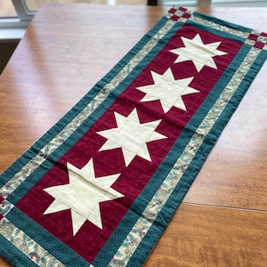 May include: A red and green table runner with a white star design. The runner has a border of green fabric with a floral pattern.