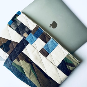 May include: A laptop sleeve made from patchwork fabric. The sleeve is made from different pieces of denim, camouflage fabric, and white fabric. The sleeve is quilted and has a diagonal pattern.