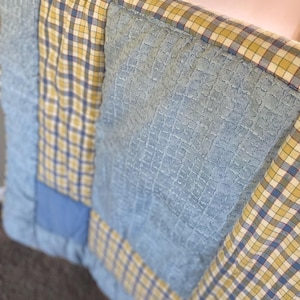 May include: Close-up of a patchwork quilt with a mix of blue, yellow, and white checkered and solid fabric squares. The quilt has a textured, quilted surface and a soft, inviting appearance. The color palette is a mix of pastel and primary colors.