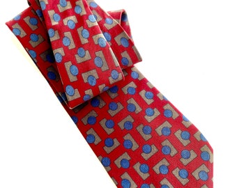 May include: A red necktie with a repeating pattern of gray squares and blue circles. The tie is knotted and lies diagonally across a white background. The fabric appears to be silk or a similar material.