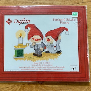May include: A Duftin Patches & Stitches cross-stitch kit featuring two gnomes in red hats, one standing by a candle on a green spool, the other beside them. The kit includes white 16ct aida, cotton floss, needle, instructions, and a red paper frame.