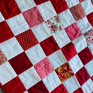 May include: A patchwork quilt featuring a checkerboard pattern of alternating white squares and various red squares. The red squares include solid colors, polka dots, floral prints, and geometric patterns, all stitched together in a grid.