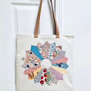 May include: A white canvas tote bag with brown straps and a colourful patchwork flower design. The flower is made of various fabrics with floral, geometric, and abstract patterns.