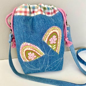 May include: Blue denim drawstring bag with floral applique. The bag features a gingham top, pink rope drawstring and strap, and decorative pink tassels. The floral applique includes pink flowers and green and yellow accents.
