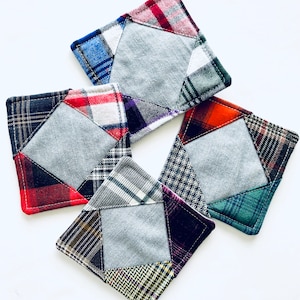 May include: Four square fabric coasters with a patchwork design. Each coaster features a gray center square surrounded by different plaid patterns in red, blue, green, and black.