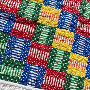 May include: A colorful patchwork quilt with a white ruffled edge. The quilt features squares in red, blue, green, and yellow, with patterns including polka dots and stripes. The quilt is a handmade item.