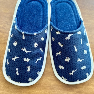 Pu&ograve; includere: Un paio di pantofole da interno blu con bordo bianco e un motivo di conigli bianchi su sfondo blu.