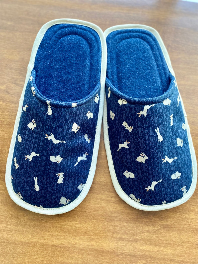 Pu&ograve; includere: Un paio di pantofole da interno blu con bordo bianco e un motivo di conigli bianchi su sfondo blu.