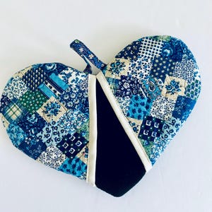May include: Heart-shaped quilted oven mitt with a patchwork design in shades of blue, green, and cream. Features a black interior and a loop for hanging. The mitt is made of fabric.