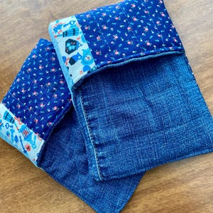 Puede incluir: Dos bolsas de mezclilla hechas a mano. Las bolsas están hechas de mezclilla azul y presentan una tela estampada floral en la solapa superior. La tela floral tiene un fondo azul con pequeñas flores rosas. Una tira decorativa con llaves y relojes adorna la parte superior.