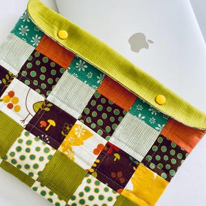 Op de afbeelding: Een patchwork laptophoes met een groene, oranje, bruine en witte kleurstelling. De hoes heeft een klep met twee gele knopen.