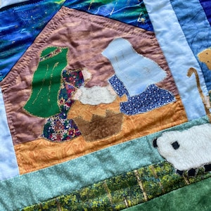 May include: A handmade quilt featuring a nativity scene. The quilt depicts Mary and Joseph with the baby Jesus in a manger. The quilt has a variety of fabrics, including blue, green, brown, and gold. A white sheep is also visible.
