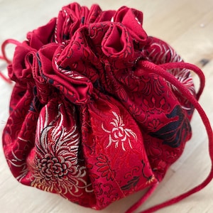 Puede incluir: Una bolsa roja con cordón y un intrincado diseño floral en blanco y dorado. La bolsa está hecha de una tela sedosa y tiene una parte superior fruncida. El cordón es rojo.