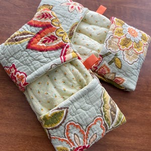 Könnte beinhalten: Zwei gesteppte Topflappen mit Blumenmustern in Orange, Rot, Gelb und Grün auf hellblauem Hintergrund. Jeder Topflappen hat eine orangefarbene Schlaufe zum Aufhängen. Der Innenstoff ist hellgelb mit kleinen floralen Details.