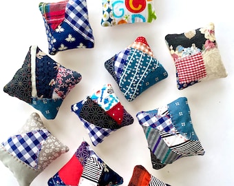 May include: A collection of ten small, square, stuffed fabric cushions featuring various patchwork designs. The textiles include blue gingham, floral prints, alphabet letters, and geometric patterns, all joined with visible hand-stitched seams.