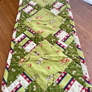 May include: A green and red quilted table runner with a patchwork design featuring a repeating pattern of mice playing musical instruments and holly sprigs.