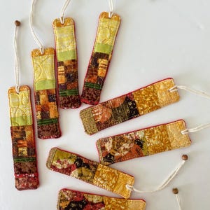 May include: A collection of handmade fabric bookmarks. Each bookmark features a patchwork design with various colors including yellow, green, red, and brown. They have a rectangular shape with a white string and a wooden bead attached.