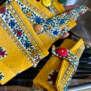 Puede incluir: Protectores de asas de ollas y agarraderas con estampado floral y amarillo. Los protectores tienen una base amarilla con un borde floral blanco y se aseguran con botones de colores. Las agarraderas tienen un diseño similar.