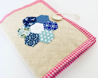 Puede incluir: Una pequeña bolsa de tela beige con un ribete de cuadros rosa y blanco. La bolsa tiene un cierre de botón y un diseño de flor de patchwork azul y blanco en la parte delantera.