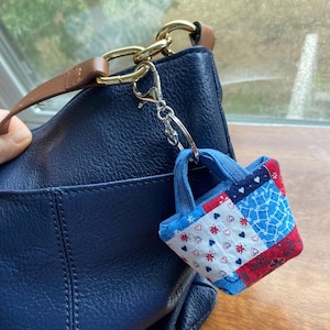 Könnte beinhalten: Ein kleiner Stoff-Tote-Bag-Anhänger mit einem Patchwork-Design in Rot, Weiß und Blau. Der Anhänger ist an einer marineblauen Handtasche mit einem braunen Lederriemen und goldfarbener Hardware befestigt. Der Anhänger hat blaue Griffe.