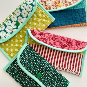 May include: A collection of handmade quilted pouches in various patterns and colors. Designs include white polka dots on green, floral prints, and red and white stripes. Each pouch has a contrasting mint green trim and a button closure.