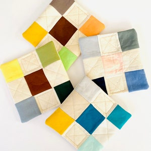 Puede incluir: Cuatro posavasos de tela acolchados con un diseño de patchwork. Cada posavasos presenta un esquema de color diferente con cuadrados de amarillo, marrón, verde, azul, naranja, gris, rosa y turquesa.