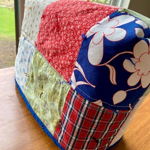 May include: A patchwork sewing machine cover featuring a variety of fabric patterns. The cover includes sections of red floral, blue floral, plaid, and other patterned fabrics. The edges are trimmed with a blue binding.