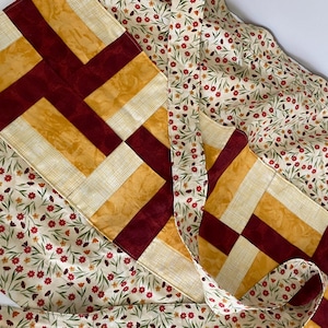 May include: A quilted table runner with a patchwork design. The runner features alternating blocks of gold, cream, and burgundy fabric. The background fabric has a cream floral pattern with red and green accents. The runner is folded and draped.