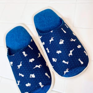 Pu&ograve; includere: Un paio di pantofole blu con un motivo di coniglio bianco. Le pantofole hanno un bordo bianco e un interno blu.