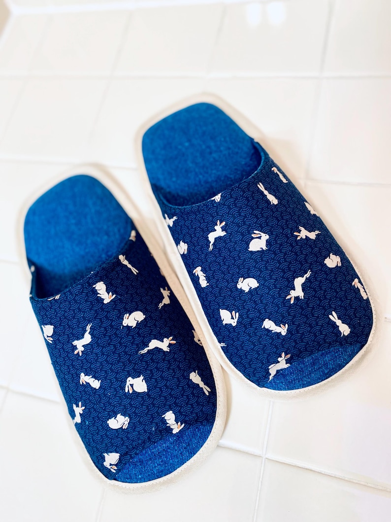 Pu&ograve; includere: Un paio di pantofole blu con un motivo di coniglio bianco. Le pantofole hanno un bordo bianco e un interno blu.