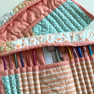 May include: A quilted fabric organizer case in peach and teal with floral and striped patterns, featuring multiple interior slots holding various colorful metal crochet hooks, a pair of silver scissors, and a white plastic needle gauge tool.