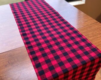 May include: Red and black checkered table runner. The runner is made of a soft, woven fabric and has a classic buffalo plaid pattern.