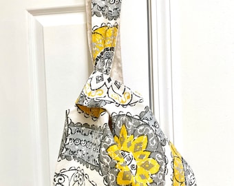 Könnte beinhalten: Eine Stofftasche mit einem Blumenmuster in Gelb, Grau und Weiß, die an einem Türgriff hängt. Das Design der Tasche zeigt große, stilisierte Blumen und geometrische Formen. Der Träger ist eine Fortsetzung des Stoffes der Tasche.