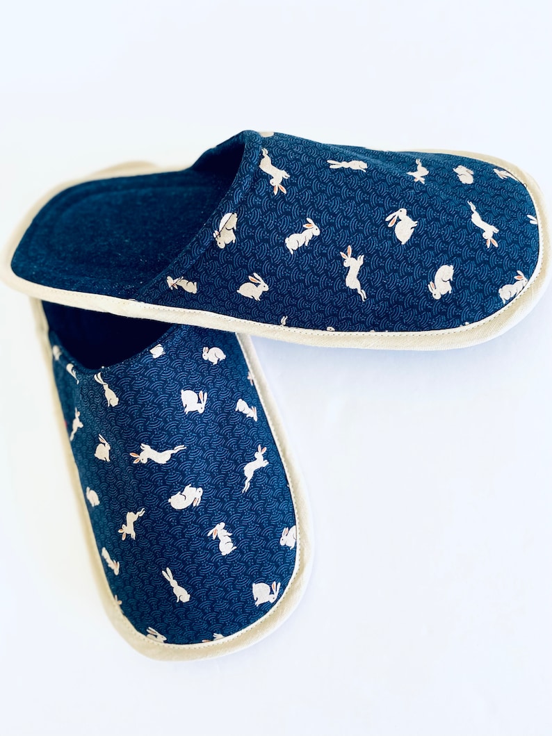 Pu&ograve; includere: Un paio di pantofole blu con un motivo di coniglio bianco. Le pantofole hanno un bordo color crema.