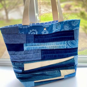 May include: A blue denim tote bag with a patchwork design. The bag has a white canvas strap and a blue paisley fabric lining.