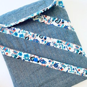 Puede incluir: Bolso de tela vaquera con rayas diagonales estampadas. Las rayas son blancas con un diseño floral azul y rojo. El bolso tiene un cierre de botón azul y un interior blanco.