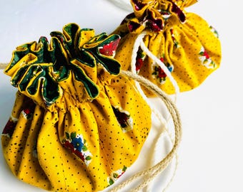 Pode incluir: Duas bolsas amarelas de tecido com cordão, padrão floral e bolinhas pretas. As bolsas têm bordas com babados com acabamento verde e bordô. Cada bolsa tem um cordão com uma conta na extremidade.