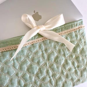Puede incluir: Una funda acolchada para portátil de color verde claro con una cinta crema atada con un lazo. La funda tiene un patrón floral y está adornada con encaje. El logotipo de Apple es visible.