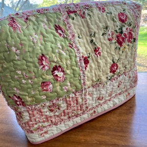 May include: A quilted tea cozy with a floral pattern. The cozy features a patchwork design with green and cream panels adorned with pink roses. The edges are trimmed with ruffled fabric and a pink border, creating a vintage aesthetic.