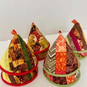 May include: Four cone-shaped quilted pot holders with hanging loops. Each has a unique patchwork design in red, orange, green, and brown fabrics. The edges are trimmed with red or green binding. A practical kitchen accessory.